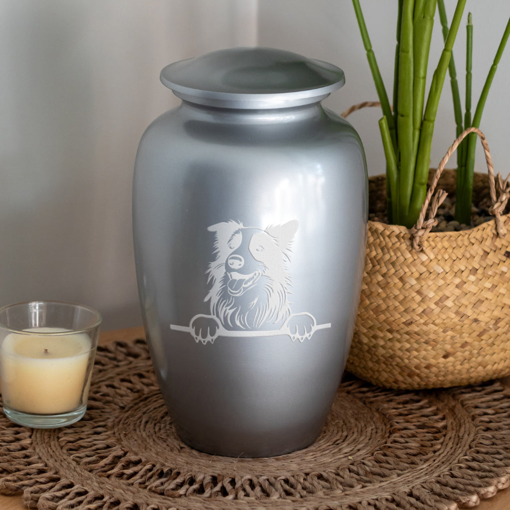 A metallic silver pet urn with a lid, featuring a white engraved design of a dog peeking over a rail, displayed on a woven mat next to a candle and a potted plant.